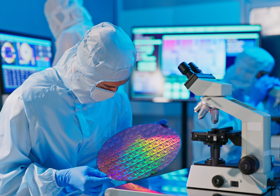 asian male technician in sterile coverall holds wafer that reflects many different colors with gloves and check it at semiconductor manufacturing plant